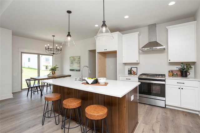 kitchen featuring wall chimney exhaust hood, a center island with sink, an inviting chandelier, stainless steel stove, and white cabinetry