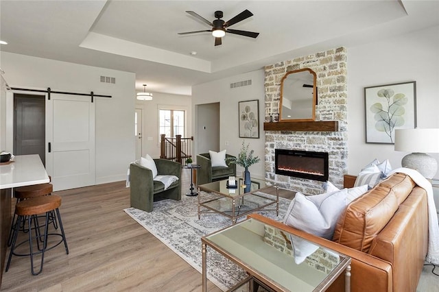 living room with ceiling fan, a barn door, a stone fireplace, light hardwood / wood-style flooring, and a tray ceiling