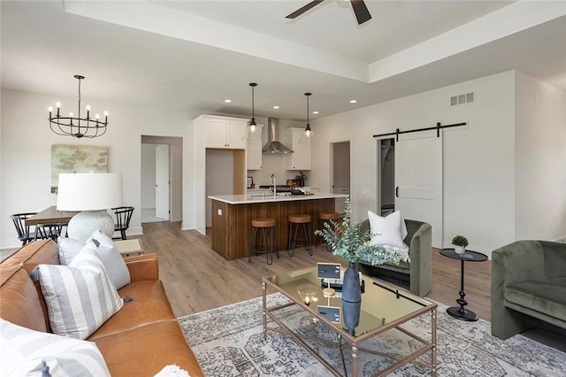 living room featuring sink, a barn door, light hardwood / wood-style floors, a tray ceiling, and ceiling fan with notable chandelier