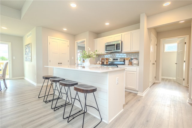 kitchen with white cabinetry, a center island with sink, stainless steel appliances, decorative backsplash, and light hardwood / wood-style flooring