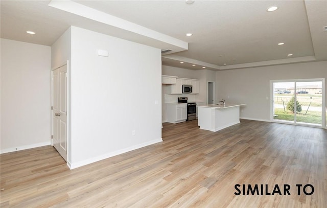 kitchen with a raised ceiling, white cabinetry, stainless steel appliances, a center island with sink, and light hardwood / wood-style flooring