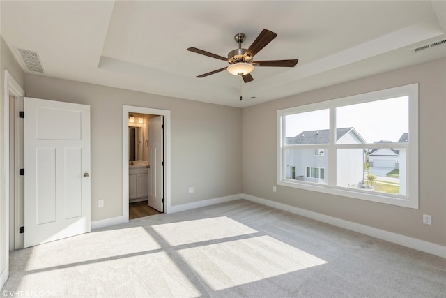 unfurnished bedroom featuring ceiling fan, a raised ceiling, ensuite bathroom, and light carpet