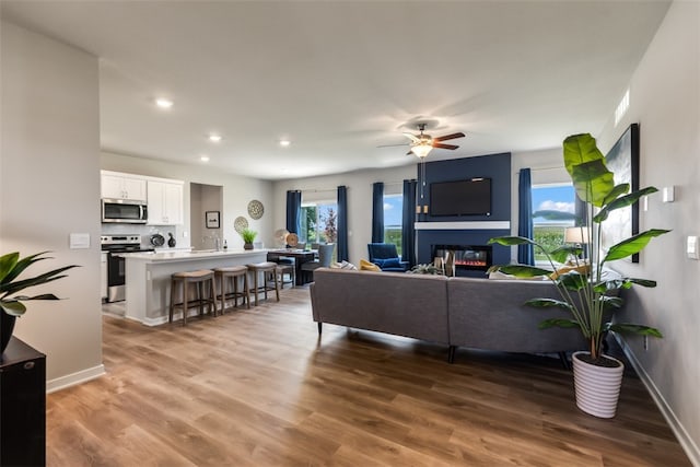 living room featuring baseboards, light wood-type flooring, recessed lighting, a glass covered fireplace, and a ceiling fan