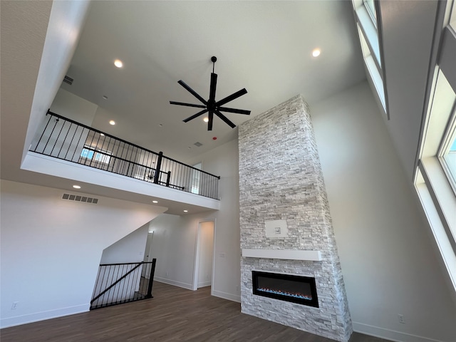 unfurnished living room with ceiling fan, a towering ceiling, a stone fireplace, and dark hardwood / wood-style flooring