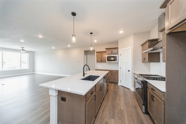 kitchen with stainless steel appliances, an island with sink, sink, hanging light fixtures, and light wood-type flooring