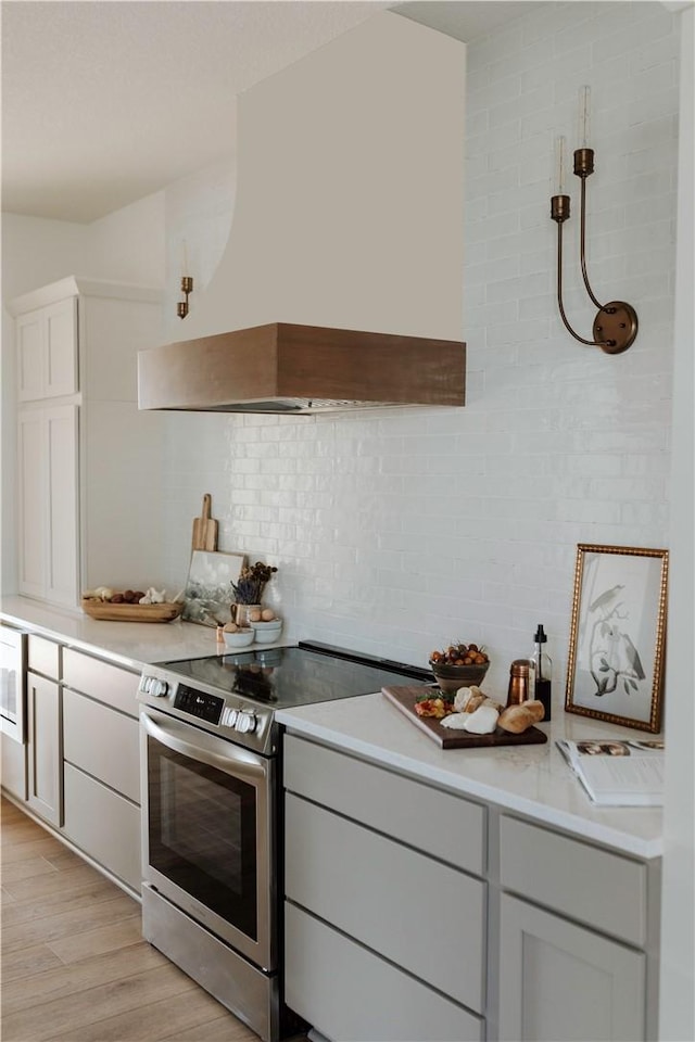 kitchen featuring tasteful backsplash, custom exhaust hood, stainless steel electric range, and light wood-type flooring