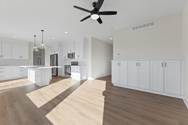 kitchen with dark wood-type flooring, appliances with stainless steel finishes, pendant lighting, a kitchen island with sink, and white cabinets