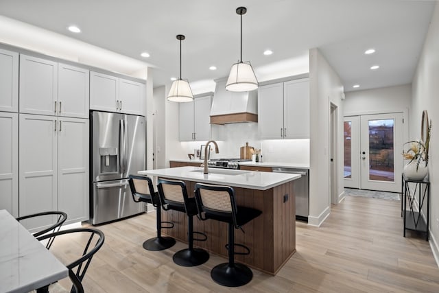 kitchen featuring appliances with stainless steel finishes, custom range hood, sink, a center island with sink, and hanging light fixtures