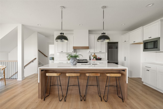 kitchen with built in microwave, white cabinetry, sink, custom exhaust hood, and a kitchen island with sink