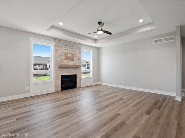 unfurnished living room with a stone fireplace, light wood-type flooring, a tray ceiling, and ceiling fan