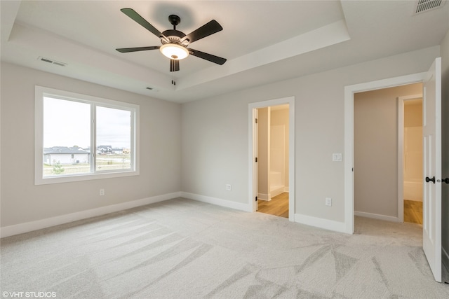 unfurnished bedroom featuring light carpet, ensuite bath, a tray ceiling, and ceiling fan