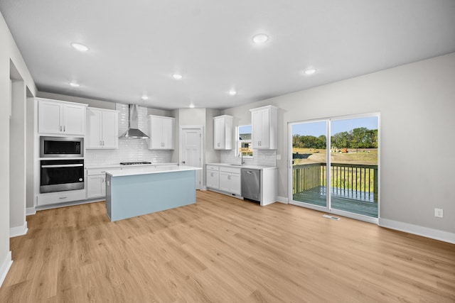 kitchen with white cabinetry, stainless steel appliances, wall chimney range hood, light hardwood / wood-style flooring, and a kitchen island