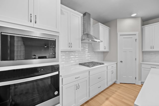 kitchen featuring white cabinetry, wall chimney exhaust hood, light hardwood / wood-style floors, decorative backsplash, and appliances with stainless steel finishes