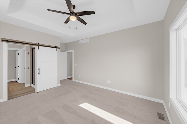 unfurnished bedroom featuring ceiling fan, a barn door, light carpet, and a tray ceiling