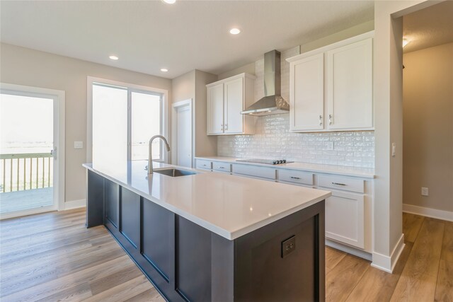 kitchen with wall chimney range hood, sink, an island with sink, white cabinets, and light hardwood / wood-style flooring