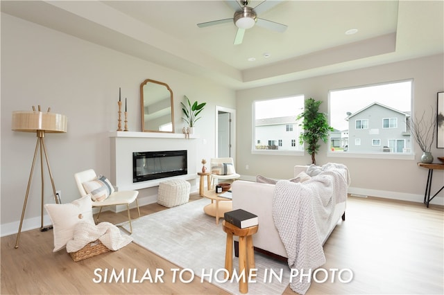 living room featuring a tray ceiling, light wood finished floors, a ceiling fan, a glass covered fireplace, and baseboards