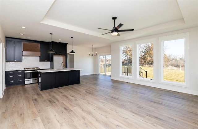 kitchen featuring an island with sink, a tray ceiling, stainless steel range, tasteful backsplash, and pendant lighting