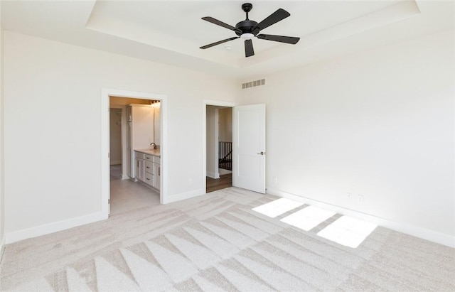 unfurnished bedroom featuring a raised ceiling, ceiling fan, light carpet, and ensuite bath