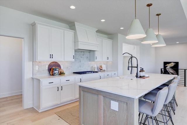 kitchen with light wood-type flooring, hanging light fixtures, white cabinetry, custom exhaust hood, and a center island with sink