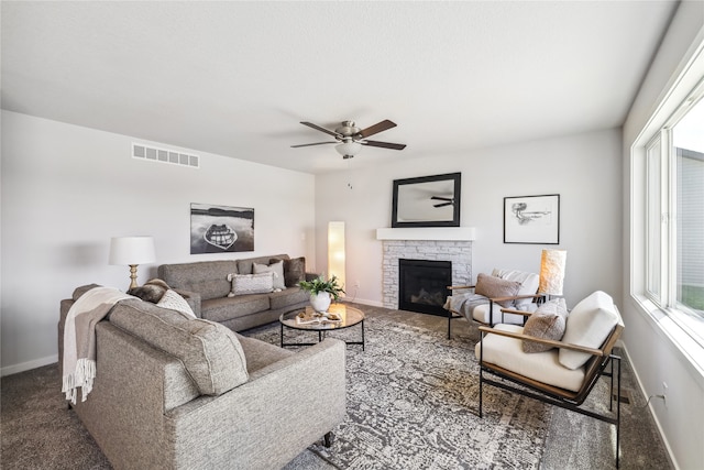 living room with a stone fireplace, ceiling fan, and dark carpet