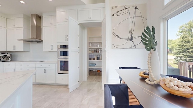 kitchen featuring wall chimney exhaust hood, light stone countertops, black electric cooktop, double oven, and white cabinetry