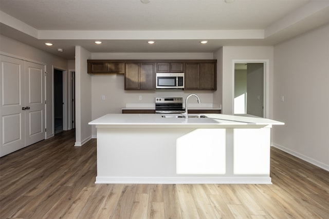 kitchen with appliances with stainless steel finishes, light wood-type flooring, dark brown cabinets, a kitchen island with sink, and sink