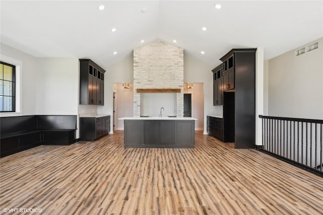 kitchen with brick wall, dark brown cabinetry, a center island with sink, hardwood / wood-style floors, and high vaulted ceiling