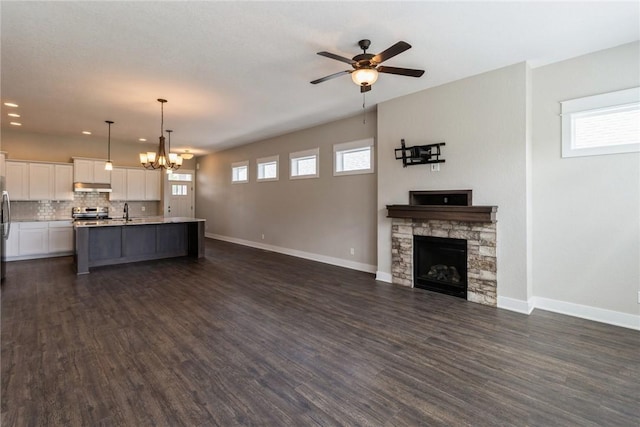 unfurnished living room featuring ceiling fan with notable chandelier, a healthy amount of sunlight, dark wood-type flooring, sink, and a stone fireplace