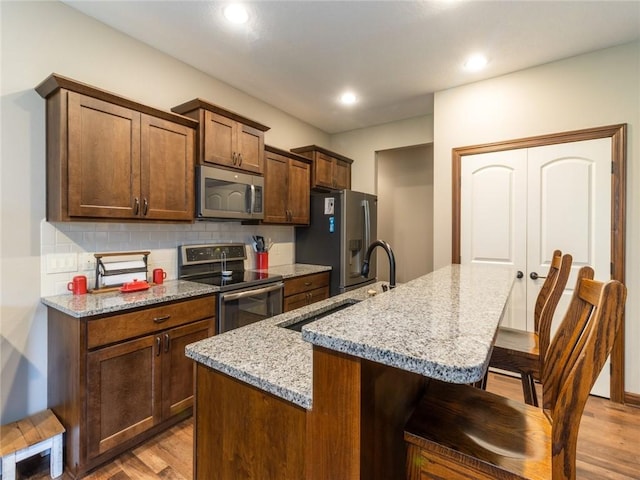 kitchen with sink, stainless steel appliances, light stone countertops, an island with sink, and light wood-type flooring