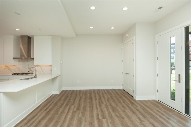 interior space with white cabinetry, sink, wall chimney range hood, light hardwood / wood-style floors, and decorative backsplash