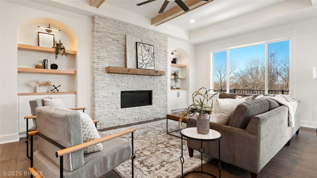 living area featuring a stone fireplace, built in shelves, dark wood-type flooring, and beam ceiling