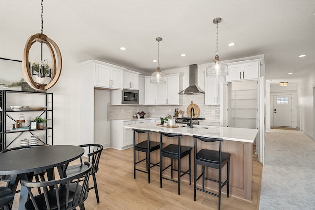 kitchen with white cabinets, wall chimney range hood, built in microwave, and an island with sink