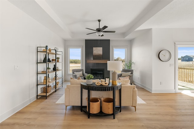 living room featuring a raised ceiling, ceiling fan, a large fireplace, and light wood-type flooring