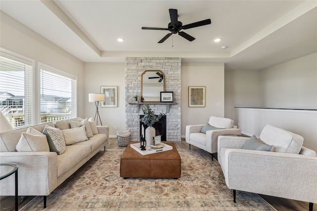 living room with a raised ceiling, wood-type flooring, a stone fireplace, and ceiling fan