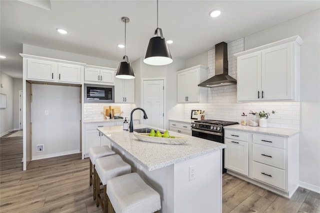 kitchen featuring built in microwave, white cabinetry, an island with sink, light stone counters, and wall chimney range hood