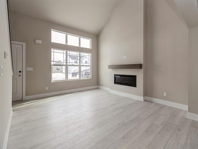 unfurnished living room with high vaulted ceiling, light wood-style flooring, baseboards, and a glass covered fireplace