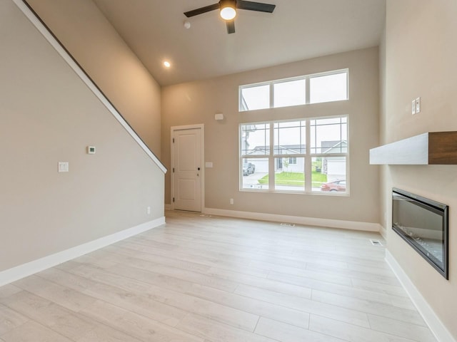 unfurnished living room featuring a ceiling fan, light wood finished floors, a glass covered fireplace, and a high ceiling