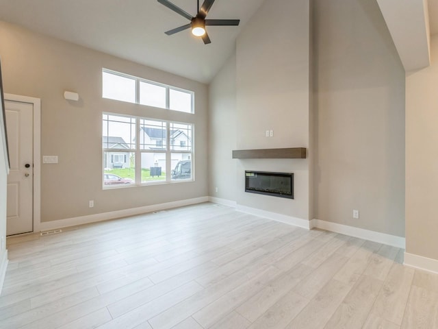 unfurnished living room featuring light wood finished floors, high vaulted ceiling, a glass covered fireplace, a ceiling fan, and a healthy amount of sunlight