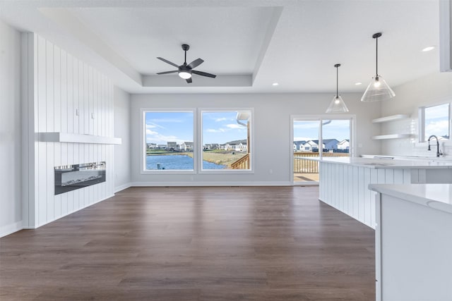 unfurnished living room featuring a water view, a tray ceiling, dark hardwood / wood-style floors, ceiling fan, and a fireplace