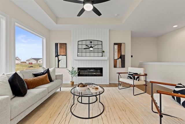 living room featuring ceiling fan, a large fireplace, a raised ceiling, and light wood-type flooring