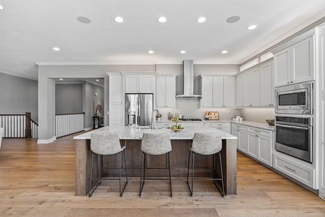 kitchen with stainless steel appliances, wall chimney range hood, white cabinets, light hardwood / wood-style floors, and an island with sink