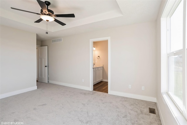 unfurnished bedroom featuring a raised ceiling, connected bathroom, ceiling fan, and dark colored carpet