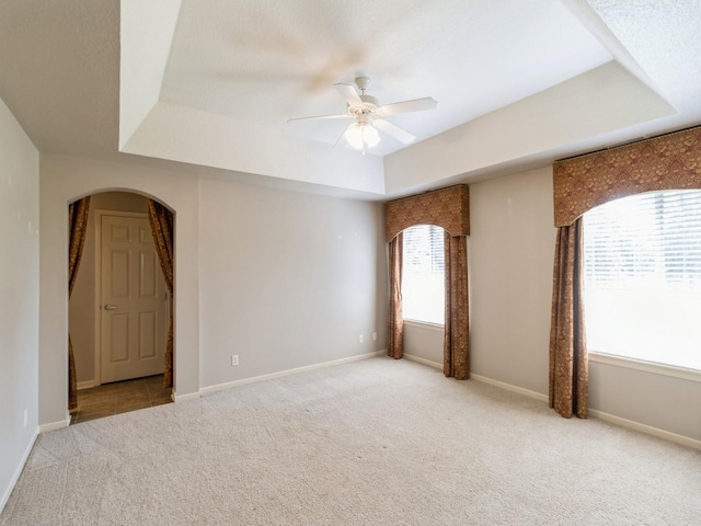 unfurnished room featuring baseboards, arched walkways, a raised ceiling, light colored carpet, and ceiling fan