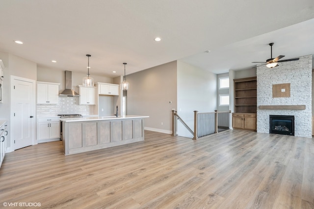 kitchen featuring wall chimney range hood, white cabinetry, a kitchen island with sink, light wood-type flooring, and a stone fireplace