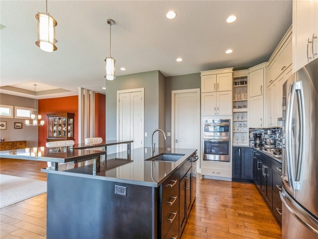 kitchen with wood-type flooring, sink, a raised ceiling, a center island with sink, and appliances with stainless steel finishes