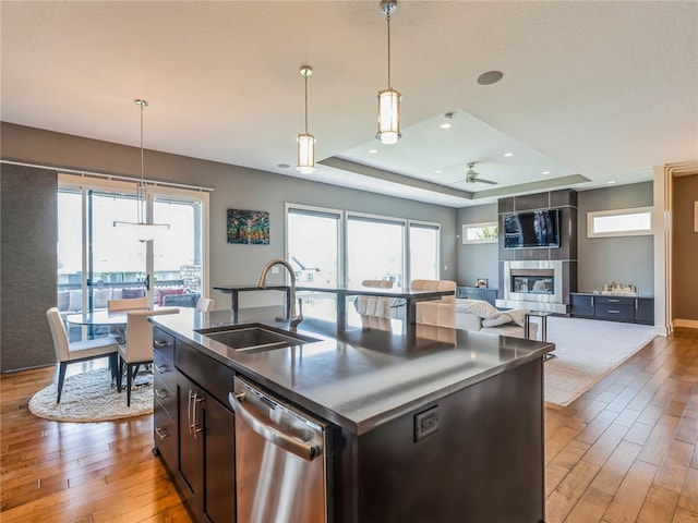 kitchen with stainless steel dishwasher, an island with sink, a raised ceiling, light hardwood / wood-style flooring, and a tile fireplace