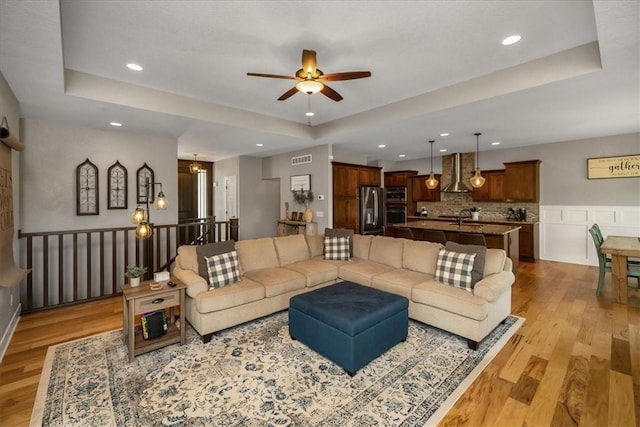 living room with sink, ceiling fan, a raised ceiling, and light wood-type flooring