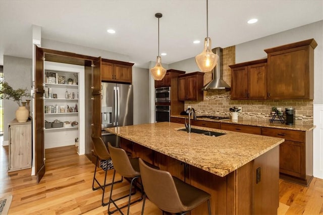 kitchen with appliances with stainless steel finishes, light hardwood / wood-style floors, a kitchen island with sink, wall chimney range hood, and pendant lighting