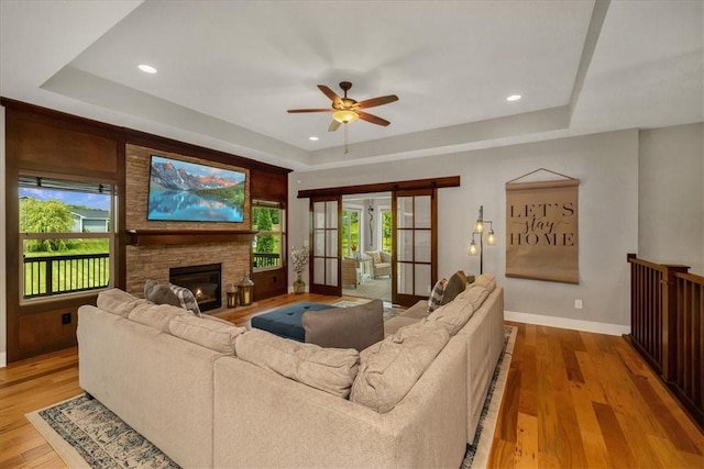 living room featuring light wood-type flooring, a fireplace, ceiling fan, and a tray ceiling