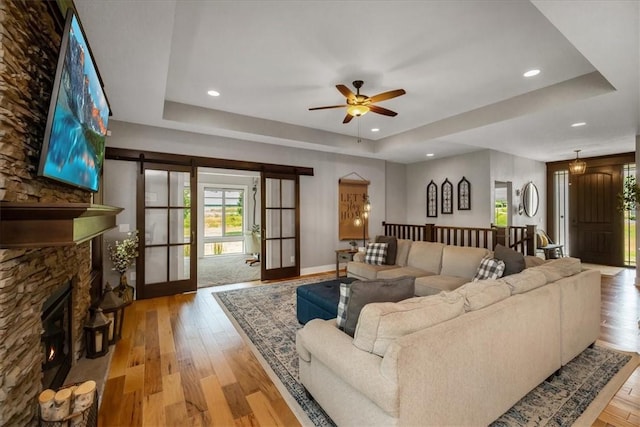 living room with ceiling fan, a stone fireplace, light hardwood / wood-style flooring, and a tray ceiling